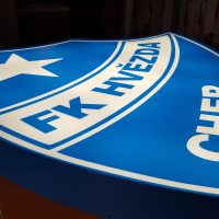 Modrý světelný panel tvaru erbu, s bílými nápisy a detaily ohledně klubu shora, vyfocený ležící z levého boku, na hnědém podkladu