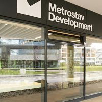 Bílé logo Metrostav Development na černém plášti nad skleněnou stěnou, výhled ven z budovy