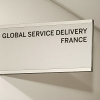 Šedá výstrč s černým nápisem GLOBAL SERVICE DELIVERY FRANCE, plexi v eloxovaném hliníkovém rámu, bílá zeď