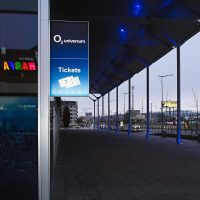 Světelná reklamní výstrč na rohu budovy O2 Universum, nápis a piktogram Tickets, skleněná stěna s odrazem loga Galerie Harfa