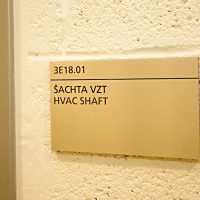 Hliníková dveřní cedulka s černým gravírovaným označením 3E18.01 a textem ŠACHTA VZT a HVAC SHAFT, bílá strukturovaná zeď