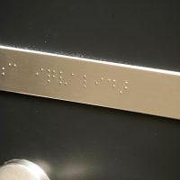 Hliníková obdélníková destička s Brailleovým písmem pro nevidomé s nápisem WC invalidé na černých dveřích