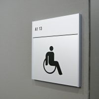 Stříbrná dveřní tabulka s označením WC pro handicapované, černý symbol osoby na invalidním vozíku, značení místnosti A1 13