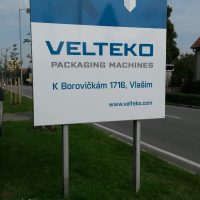 Reklamní cedule firmy VELTEKO s šipkou přesahující okraj, stojící na dvou sloupech v exteriéru na trávníku, v pozadí část silnice, stromy, obloha