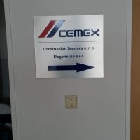 Čtvercová stříbrná reklamní deska s logem CEMEX, informačními nápisy a šipkou doprava, nalepená na bílé stěně mezi futry, pod ní vypínač