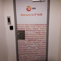 Dveře na chodbě polepené fototapetou s logem NN, sloganem a oranžovými nápisy ve světových jazycích