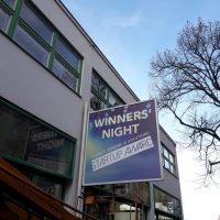 Velká reklamní výstrč na fasádě dvou podlažní budovy, nápis WINNERS‘ NIGHT, řady oken a výloh, pojízdná montážní plošina