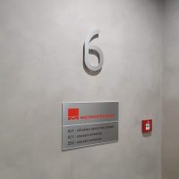 Bílá plastická číslovka 6 na zdi jako označení šestého patra, připevněná distančně na zdi, pod ní kovová informační tabulka s červeným logem a černým textem
