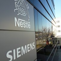 Dvě jemná bílá loga firem Nestlé a Siemens umístěná na šedém plášti budovy v blízkosti zrcadlové černé plochy
