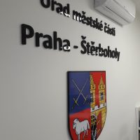 Černý 3D nápis Úřad městské části Praha - Štěrboholy níže doplněný barevným erbem přilepený na bílou zeď, pod klimatizační jednotku