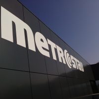 Detailněji vyfocené bílé logo firmy metrostav přilepené na černém plášti budovy v pozadí s modrou oblohou.