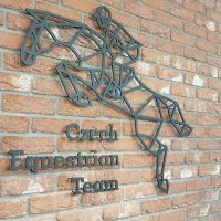 Černé geometrické logo ve tvaru jezdce na koni doplněné o černý nápis Czech Equestrian Team připevněné na cihlovou zeď v blízkosti černých otevřených dveří