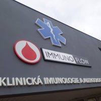 3D modro-bílé logo záchranné služby, níže bílo-červené logo kapky krve, vpravo černo-bílé logo s nápisem immunoflow, dole bílý 3D nápis klinická imunologie a alergologie, přidělané na šedou fasádu budovy