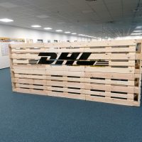 Černý nápis/logo firmy DHL nalepený na stěnu z dřevěných palet v exteriéru s modrým kobercem.