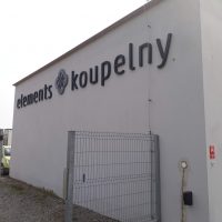 Černý 3D nápis a černé 3D malé logo ve tvaru čtyřlístku přilepené na budovu tvaru kvádru šedé barvy do horní části. V popředí vstupní branka a v pozadí auta.