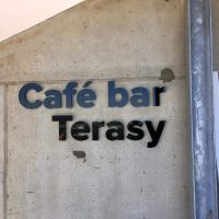 Černomodrý 3D nápis café bar Terasy umístěný na šedou betonovou stěnu.