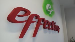 Mohutný 3D červený nápis effective nahoře doplněný o bílé 3D logo tvaru e na světle zeleném nepravidelně kruhovém podkladu, umístěno na bílé stěně v interiéru