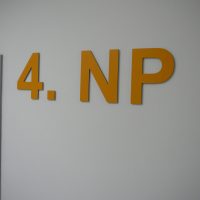 Žlutý plastický 3D nápis 4. NP na bílé zdi vedle šedého rámu dveří, označení čtvrtého podlaží budovy