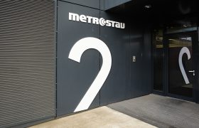Označení budovy částí číslice 2 z bílého dibondu na černém plášti budovy, bílé logo Metrostav, odraz ve skleněných dveřích za čistící zónou