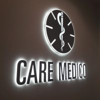 Tmavě šedý nápis CARE MEDICO a nad ním kulaté logo, podsvíceno bílým světlem, umístěno na světlé stěně