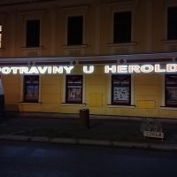 Ve tmě vyfocený žlutý dům s klasicistní fasádou, dvě řady oken, dolní řada oken potištěná reklamami, nad okny nápis POTRAVINY U HEROLDŮ, oranžový s bílým obrysem, svítící, nad tím přerušovaná tmavší čára na bílém podkladu přes celý dům, v popředí asfalt a cyklo stojan