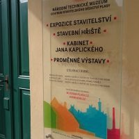 Skleněná deska na pískově žluté fasádě, označení expozice, Národní technické muzeum, barevné siluety budov, nerezové distance