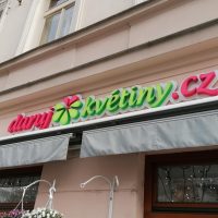 Detailně vyfocený panelový dům, ve středu logo daruj květiny.cz, červeno zelené, uprostřed květinka, nad tím další část domu, pod logem dvě vysouvací markýzy, pod markýzami okna