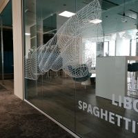 Bílé geometrické tvary a nápis JE LIBO SPAGHETTI?, inkoustová grafika nalepená na skleněné stěně moderního restauračního zařízení