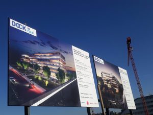 Dvě velkoplošné cedule na staveništi s vizualizací budoucích staveb administračních budov DOCK, v pozadí modré nebe a jeřáb