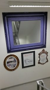 Čtvercové zrcadlo s mohutným modrým historickým rámem, pod ním vlevo šedobílý obraz ve zlatém kulatém rámu, vprostřed šedobílý obraz v černém obdélníkovém rámu a vpravo šedobílý obraz v rámu tvaru erbu, vše na bílém podkladu, v mezipatře u schodiště, které se odráží v zrcadle, nahoře zářivka