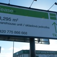 Pohled zespodu na dvojitý reklamní billboard, dibond deska s potištěnou folií, černý rám a sloup, tři světla instalovaná na vrchu. V pozadí část budovy HOPI a reklamní cedule SCANIA, obloha