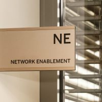 Šedá informační výstrč na zdi, černé označení NE NETWORK ENABLEMENT, skleněné okno s žaluziemi