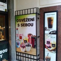 Reklama na osvěžení od MCDonalds na dibondové desce instalované na kovové mříži, vpravo reklamní fotografie produktů na desce v černém rámu instalované na zdi, vlevo část provozovny