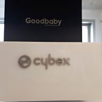 Dva nápisy z lakovaného šedého plexiskla umístěné na stěnách. Logo a nápis cybex lepené na černý hliníkový dibond umístěné na bílý podklad. V horní části šedý nápis Goodbaby na černé stěně před dvěma okny.