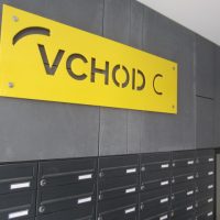 Žlutá deska s vyřezaným nápisem VCHOD C na šedém obložení zdi nad sestavou černých poštovních schránek bytového domu