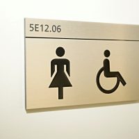 Hliníková dveřní tabulka s černým gravírovaným označením 5E12.06 a piktogramy WC ženy a invalidé, bílá strukturovaná zeď