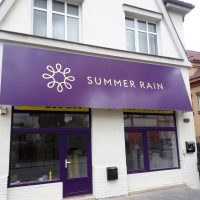 Fialová reklamní výstrč nad výlohami malého obchodu, imitace markýzy, bílé firemní logo a nápis SUMMER RAIN