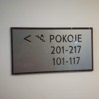 Informační skleněný štítek s bílým pozadím, černým rámečkem a gravírovaným označením pokojů, piktogramem schodů a šipkou, nalepený na bílé zdi