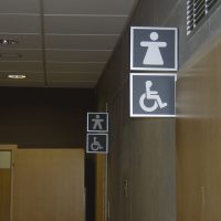 Informační výstrče označující toalety, stříbrné piktogramy panáček, panenka a vozíčkář v černé ploše, WC muži, ženy, invalidé