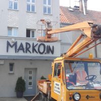 Oranžové vozidlo s vysokozdvižnou plošinou, nainstalované černé 3D logo MARKO.IN na fasádě budovy, zástavba