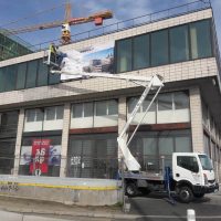 Technik na vysokozdvižné plošině instaluje potištěný banner na okna rohové budovy, v pozadí jeřáb s označením Metrostav