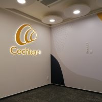 Laminová fototapeta s geometrickými tvary nalepená na stěně, svítící logo Cochlear na bílé zdi, černá podlaha, designové kruhové osvětlení