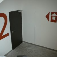 Malované označení patra na bílé zdi bytového domu, velká červená číslice 2 a směrový piktogram k výtahu, černé dveře
