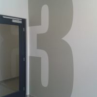 Malovaná šedá číslovka 3 přes celou výšku bílé stěny jako značení uvnitř interiéru, šedá podlaha, vlevo část prosklených dveří