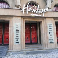 Vstup do budovy Hamleys, tři vchody, prosklené dveře s červenou grafikou, dvě informační cedule z mléčného skla na sloupech