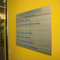 Modulová orientační tabule Ústavu molekulární genetiky AV ČR, kovová deska s modrými popisky místností, přišroubováno na žluté zdi, detail skleněných dveří