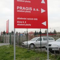 Červená cedule na šedých nohách, bílé navigační šipky a nápisy, betonové patky v trávě, dvě stříbrná auta za drátěným plotem