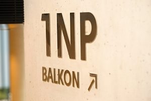 Označení patra 1 NP, text balkon a šipka, vyřezáno z nerezu, nalepeno na betonové zdi