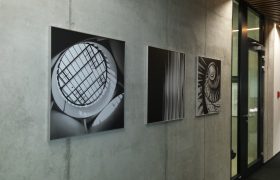 Tři černobílé fotografie jako obrazy ve stříbrných rámech na betonové stěně, v pozadí prosklené dveře s černým rámem