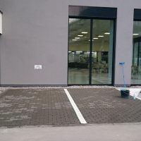 Bílé linky z epoxidových podlahových barev jako označení parkovacích míst před budovou, šedá budova s prosklenými dveřmi, nářadí potřebné pro provádění malby